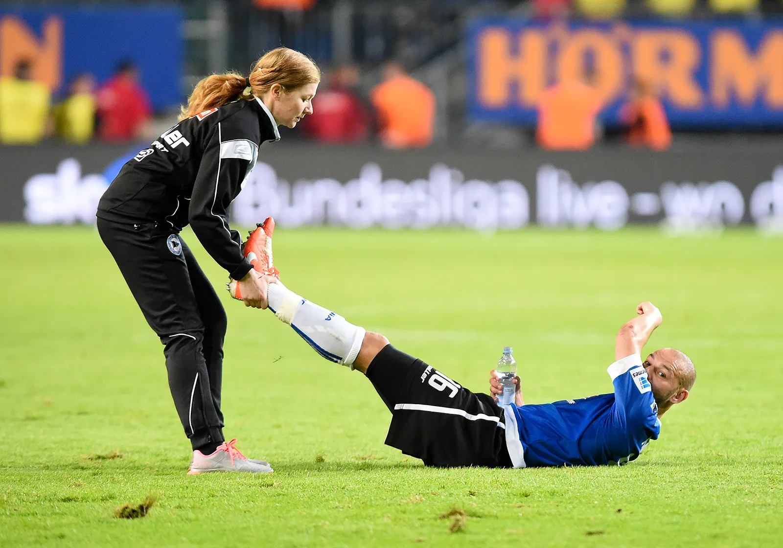  Jessica Mathiak in schwarzer Arminia Bielefeld Teamkleidung dehnt das Bein einer am Boden liegenden Spielerin in blauem Trikot während eines Fußballspiels im Stadion