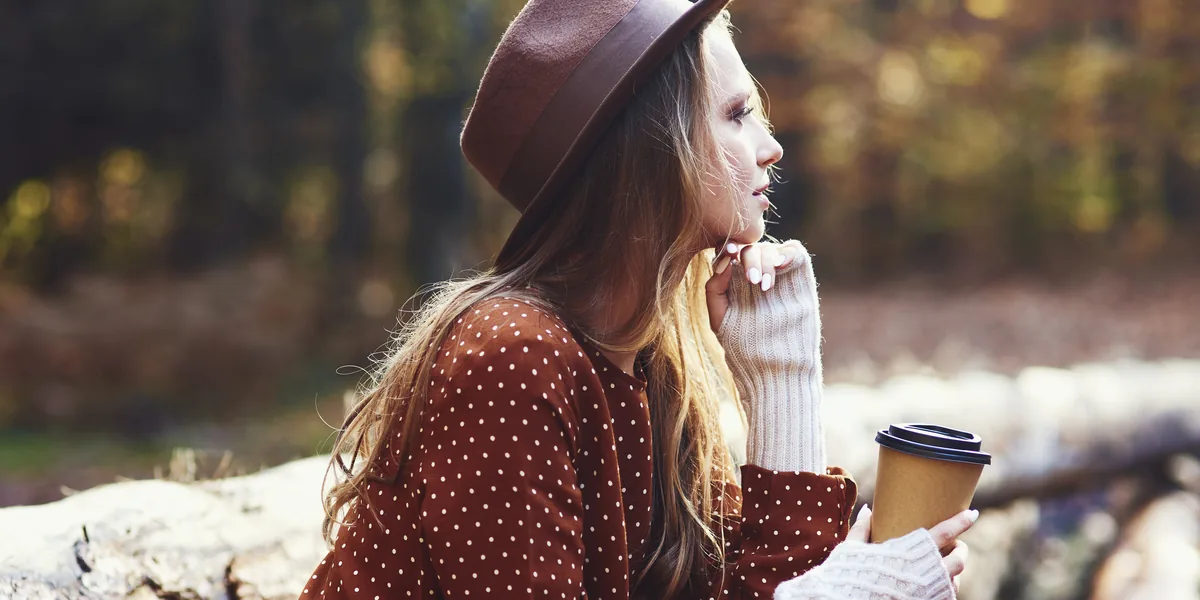 Person in a rust polka-dot dress and cream knitted arm warmers sitting on logs in an autumn wood, holding a takeaway coffee