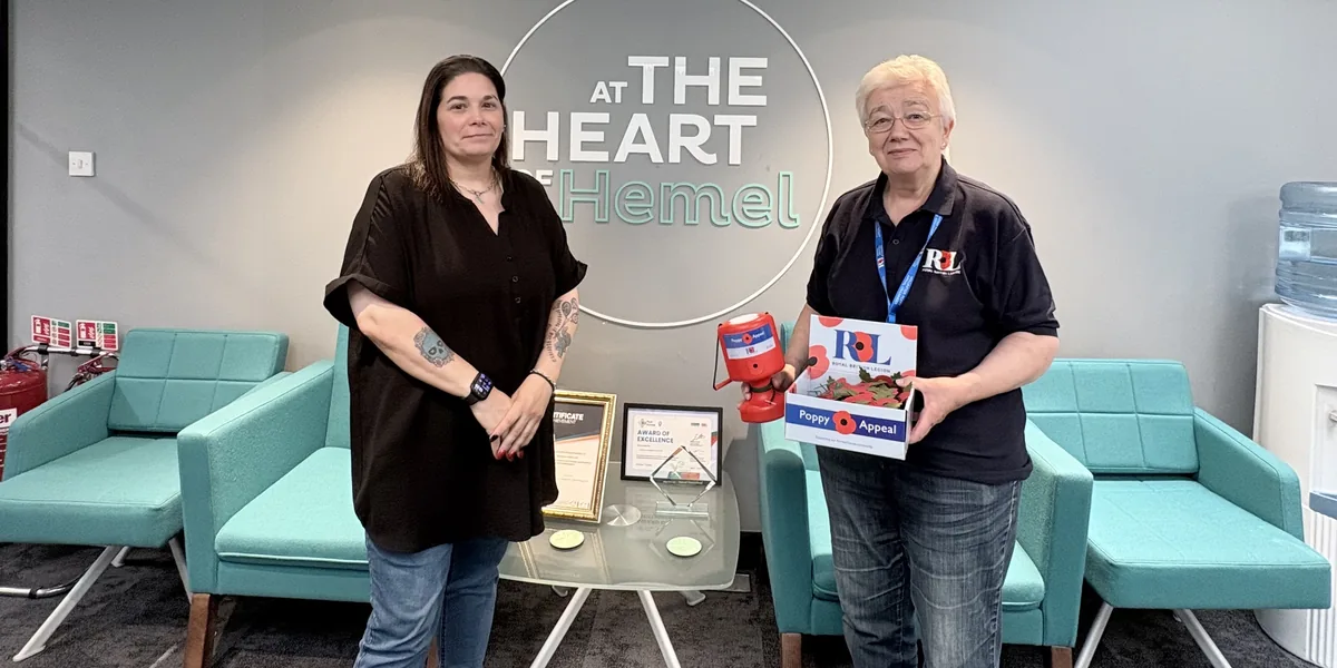 Two people standing in front of an 'At the Heart of Hemel' sign, one holding a Royal British Legion Poppy Appeal collection tin and box of poppies