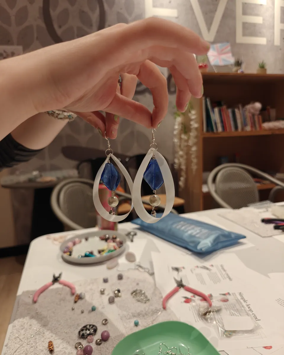 Hand holding a pair of handmade teardrop earrings with blue glass beads above a table of jewellery-making tools and beads