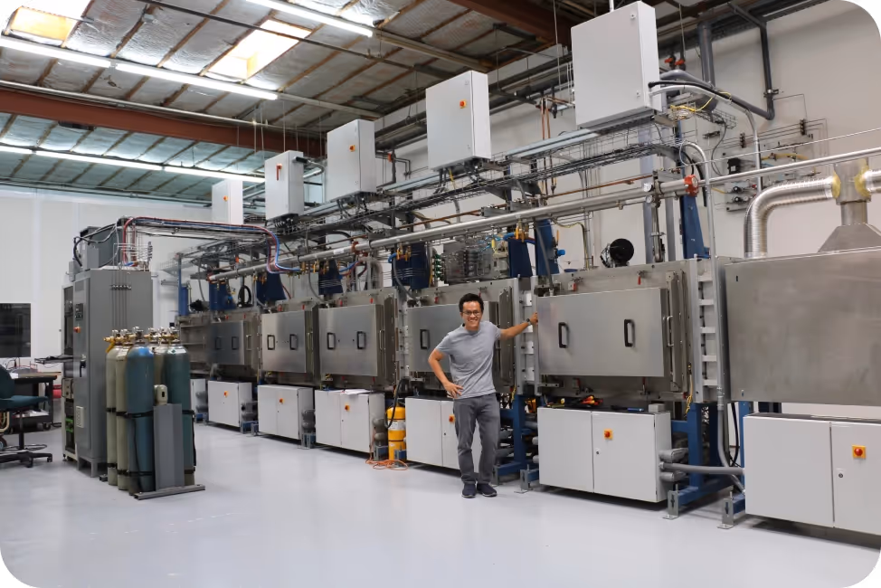 Man standing in a large industrial laboratory next to a long row of connected metal machines and gas cylinders.