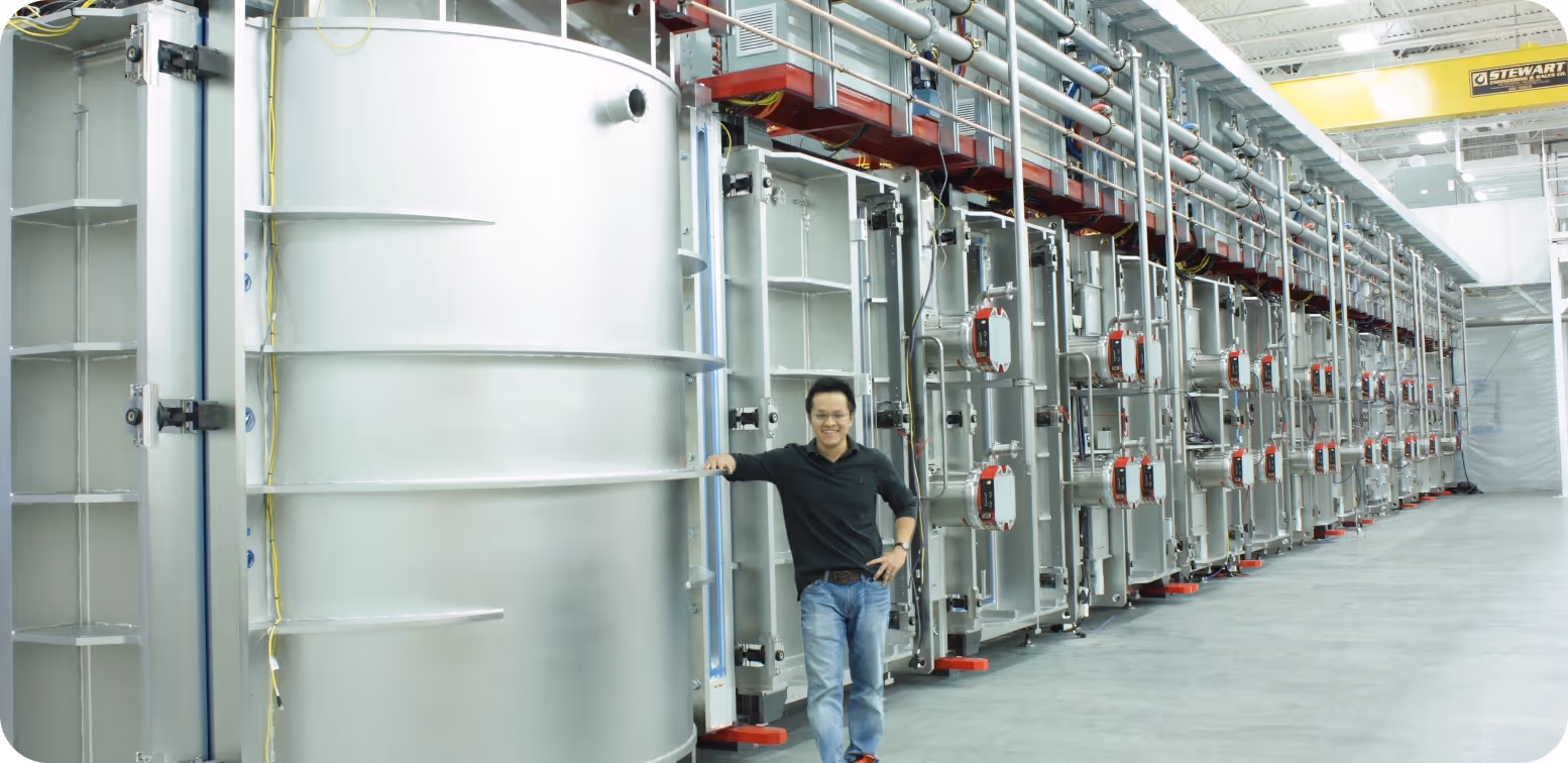 Man standing and leaning on a large industrial metal tank inside a spacious, modern factory with rows of industrial equipment.