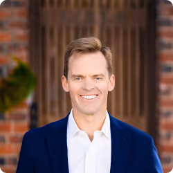 Smiling man with light brown hair wearing a white shirt and navy blazer standing in front of a brick building with a wooden door.