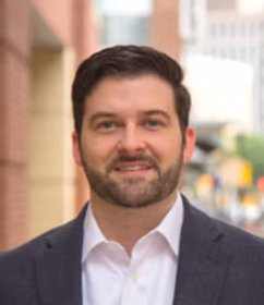 Smiling man with dark hair and beard wearing a dark blazer and white shirt, standing outdoors in an urban setting.