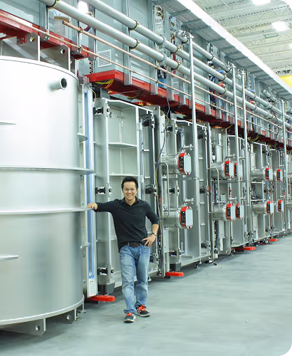 Man standing and leaning against large industrial metal tanks with pipes in a spacious factory setting.