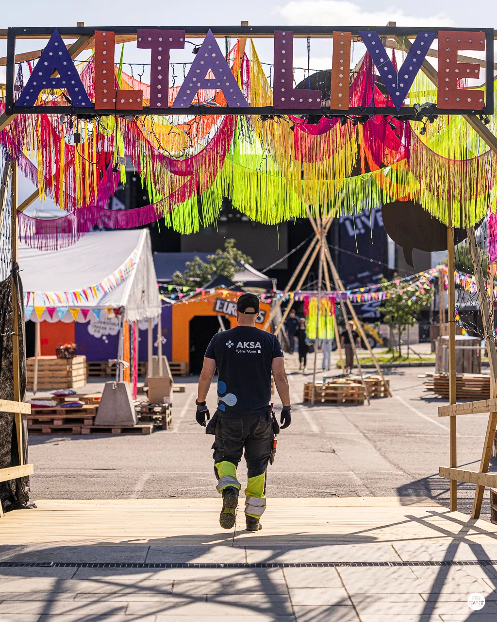 Person går under inngangsportal prydet med fargerike frynser og skiltet ALTA LIVE ved et utendørs arrangement.