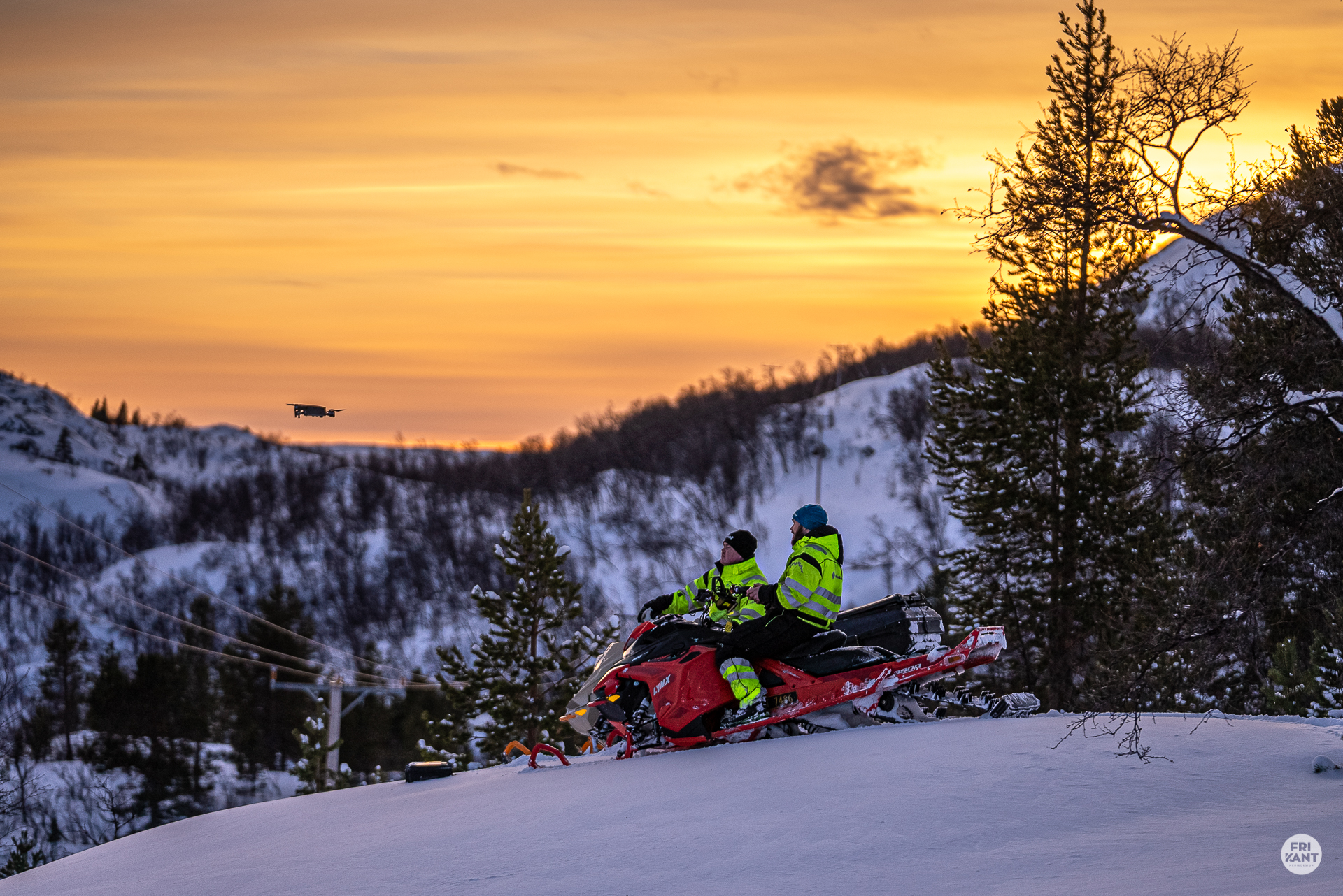 To personer i gule vinterjakker kjører snøscooter i snødekt fjelllandskap under solnedgang med en drone flyvende i bakgrunnen.