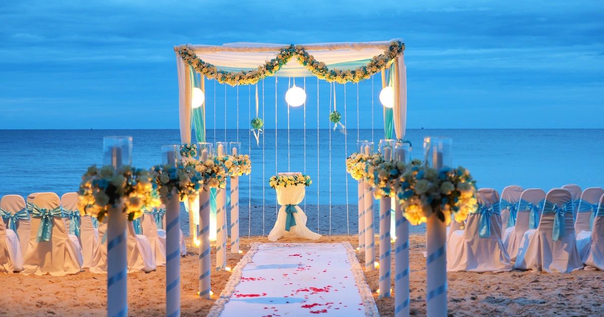 Allée de mariage décorée sur la plage au coucher du soleil avec arche fleurie et chaises blanches ornées de rubans bleus.