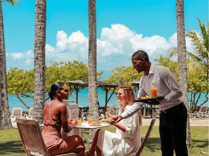 Serveur apportant des boissons à des clientes en terrasse face à la mer à l’hôtel La Créole Beach au Gosier en Guadeloupe.