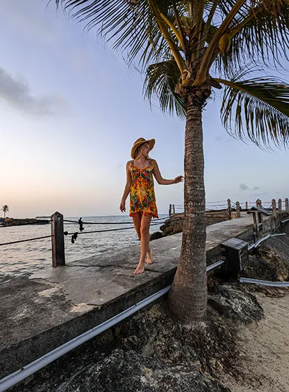Woman in a dress and straw hat walking along the Caribbean Sea in the tropical setting of La Créole Beach Hotel & Spa in Le Gosier, Guadeloupe.