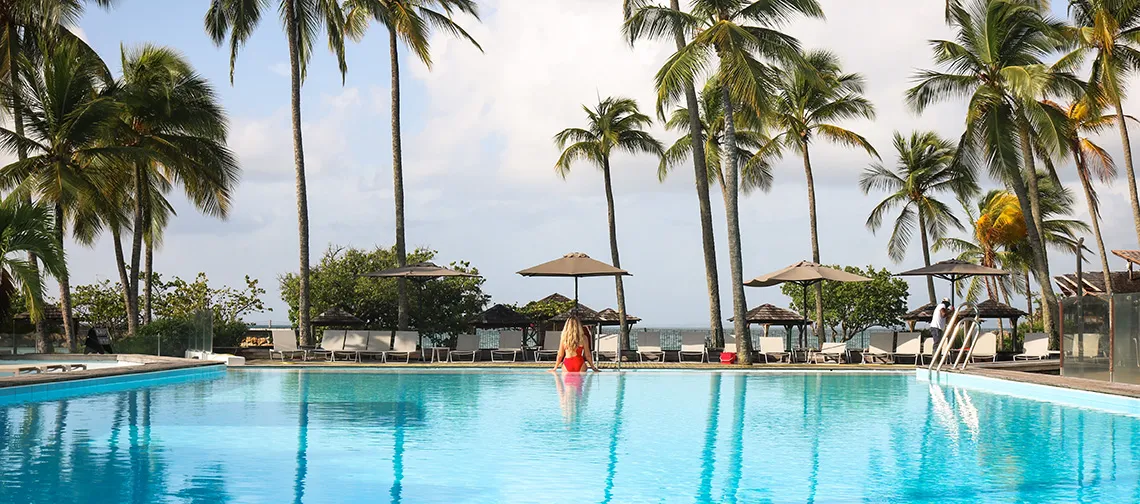 Großer tropischer Pool, umgeben von Palmen und Sonnenschirmen mit Blick auf das Karibische Meer im Luxushotel La Créole Beach Hotel & Spa in Le Gosier, Guadeloupe.