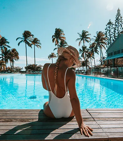 Frau in weißem einteiligem Badeanzug und Strohhut genießt den Blick auf den tropischen Pool im 4-Sterne-Hotel La Créole Beach Hotel & Spa in Le Gosier.