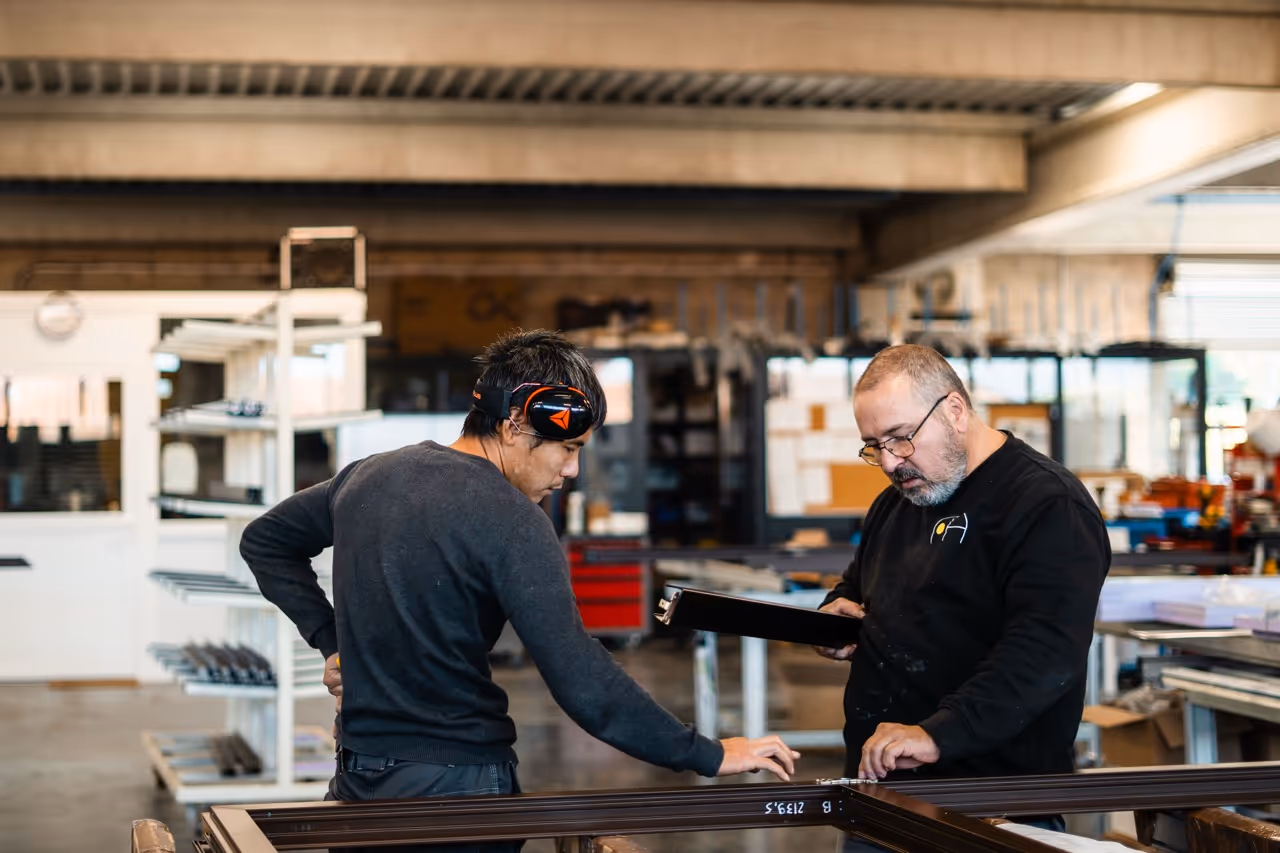 Deux ouvriers examinent un cadre en atelier industriel, l'un avec des protections auditives et l'autre tenant un dossier.