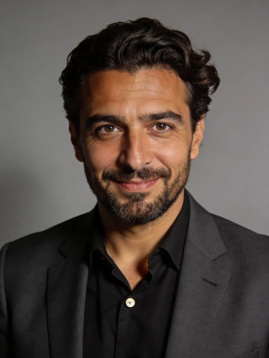 Portrait of a man with dark curly hair and beard, wearing a black shirt and dark blazer, smiling against a gray background.