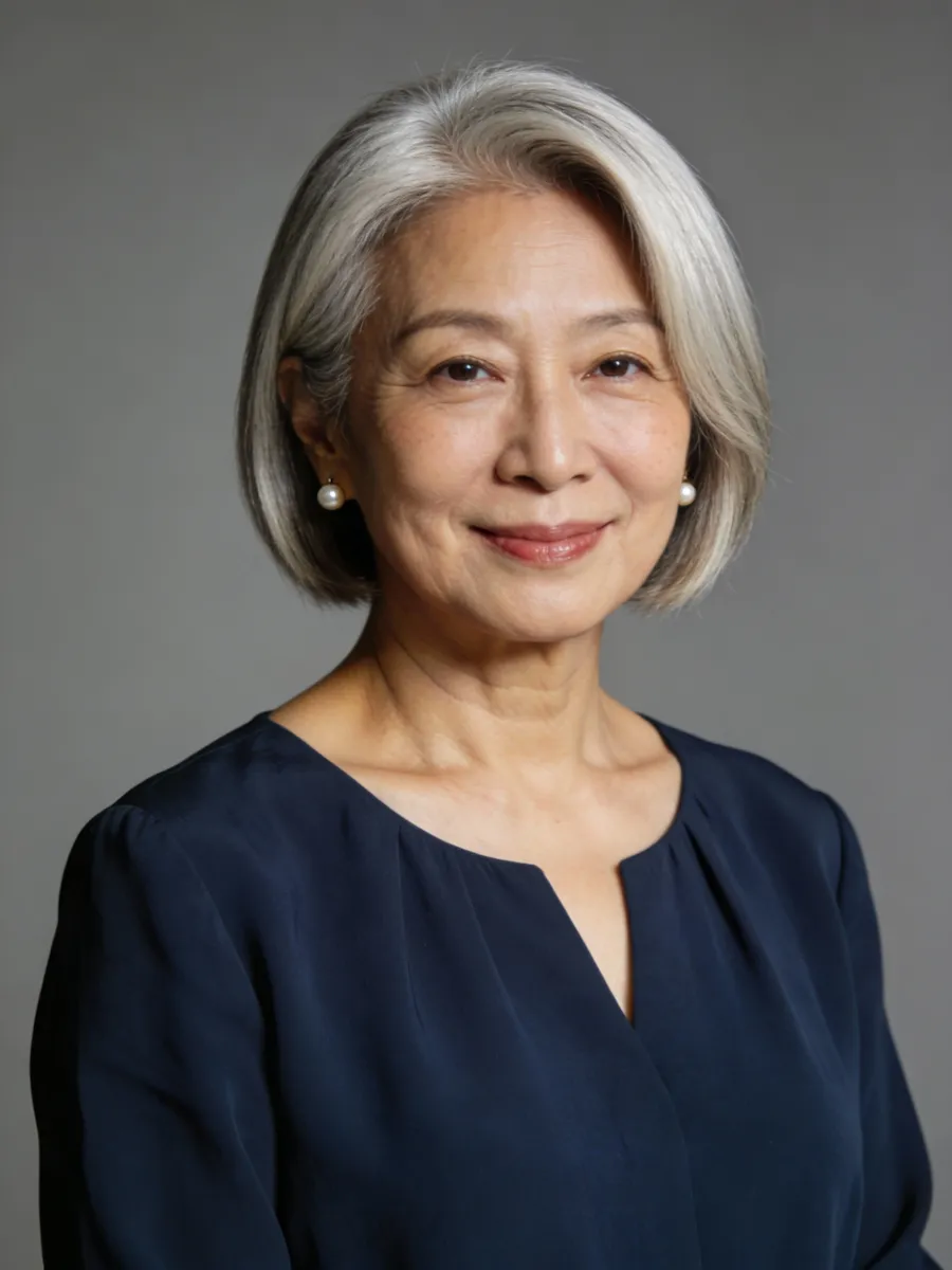 Smiling mature woman with short gray hair wearing pearl earrings and a dark blue blouse against a gray background.