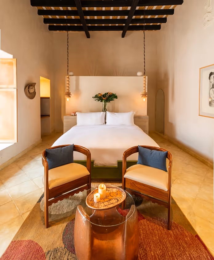 Luxury hotel suite interior featuring a king-size bed, exposed beam ceiling, two designer wooden armchairs, and soft candle lighting.