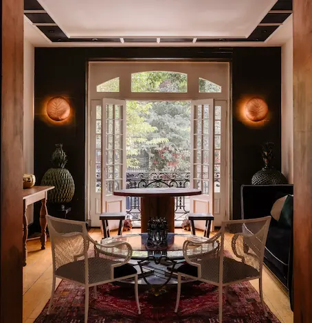 Luxury contemporary dining room featuring a circular glass table, rattan chairs, and large French doors opening onto a lush green garden.