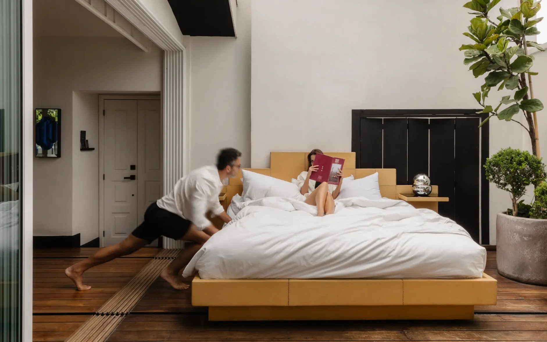 Modern minimalist bedroom featuring a couple: a woman reading a magazine in bed and a man sitting down, highlighting sleek and bright interior design.