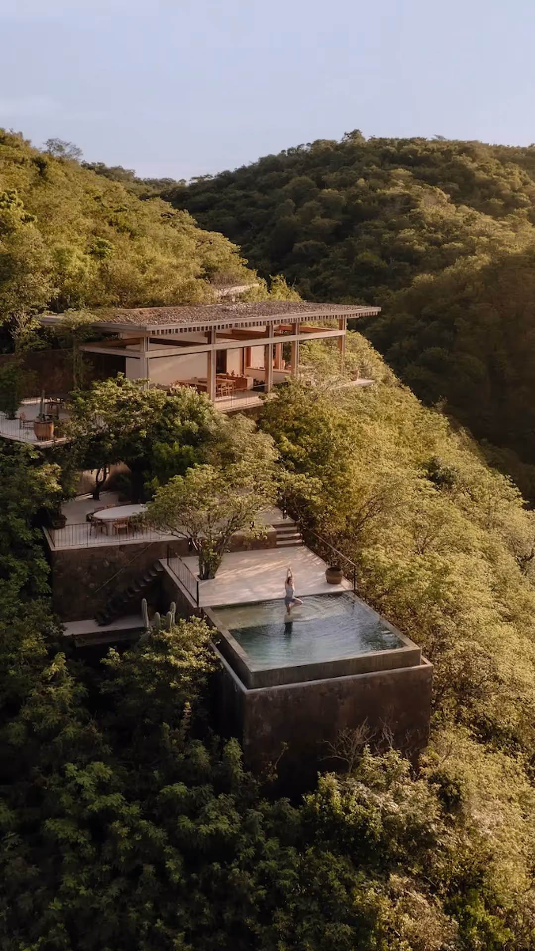 Aerial view of a modern luxury villa with brutalist architecture nestled in a lush green hillside, featuring green rooftop terraces and seamless integration into the wild landscape.