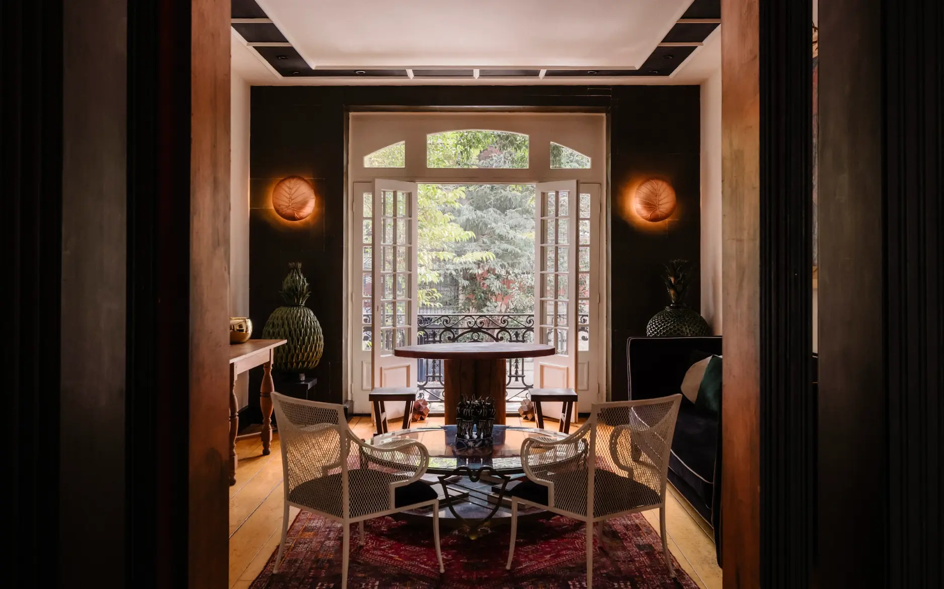 Luxury contemporary dining room featuring a circular glass table, rattan chairs, and large French doors opening onto a lush green garden.