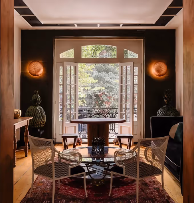 Luxury contemporary dining room featuring a circular glass table, rattan chairs, and large French doors opening onto a lush green garden.