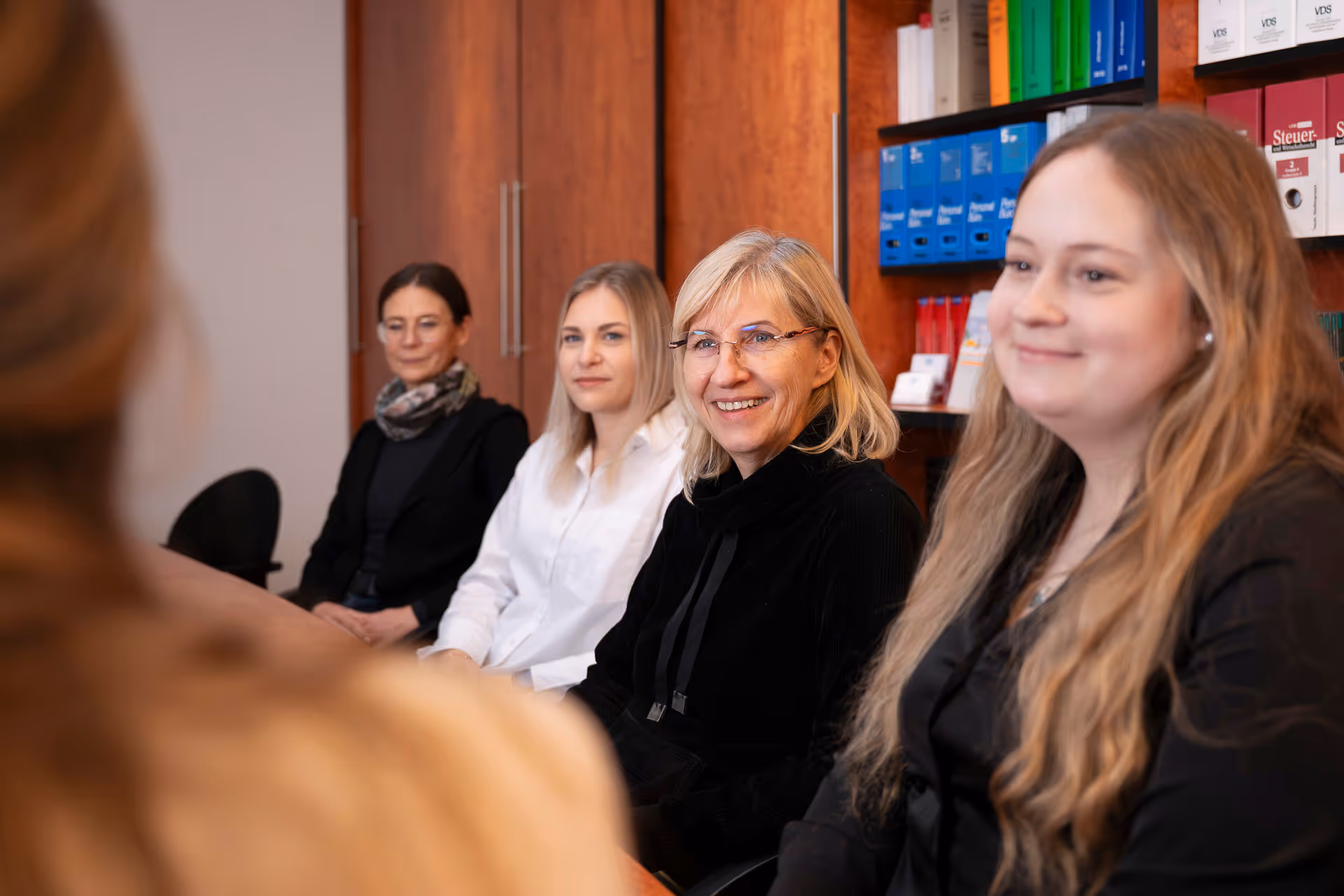 Vier Frauen sitzen an einem Holztisch im Büro, im Hintergrund Regale mit Ordnern.