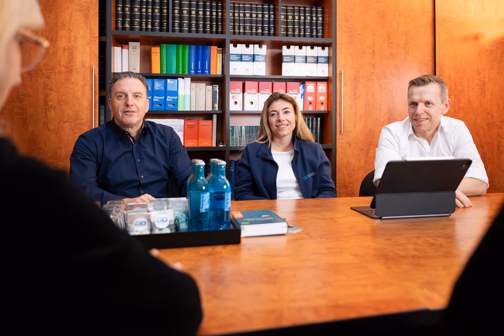 Drei Personen in einem Besprechungsraum mit Holzwänden und Bücherregal, zwei Männer und eine Frau lächeln.