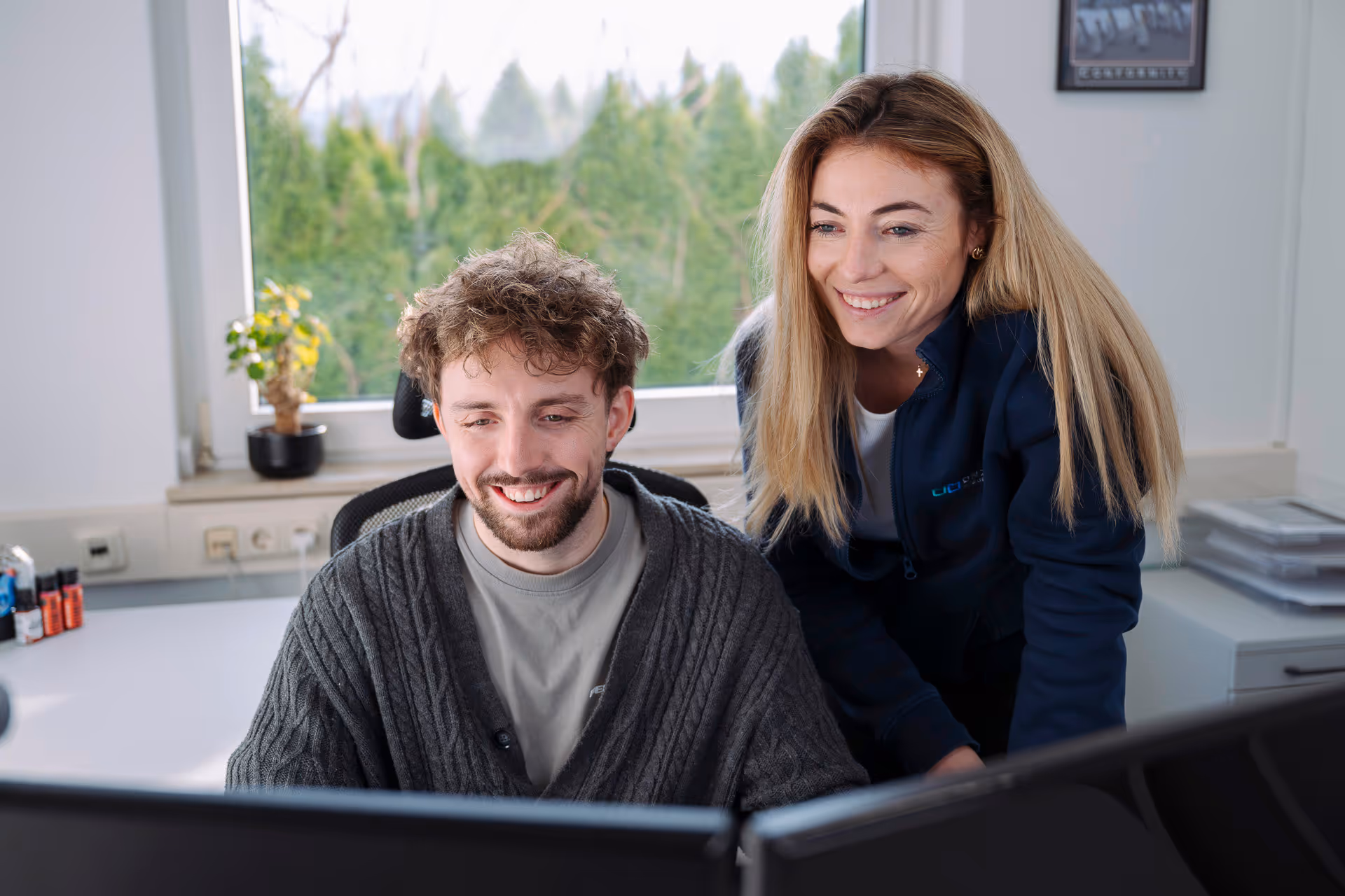 Zwei lächelnde Personen arbeiten gemeinsam an zwei Computermonitoren in einem hellen Büro mit Fenster mit Blick auf grüne Bäume.