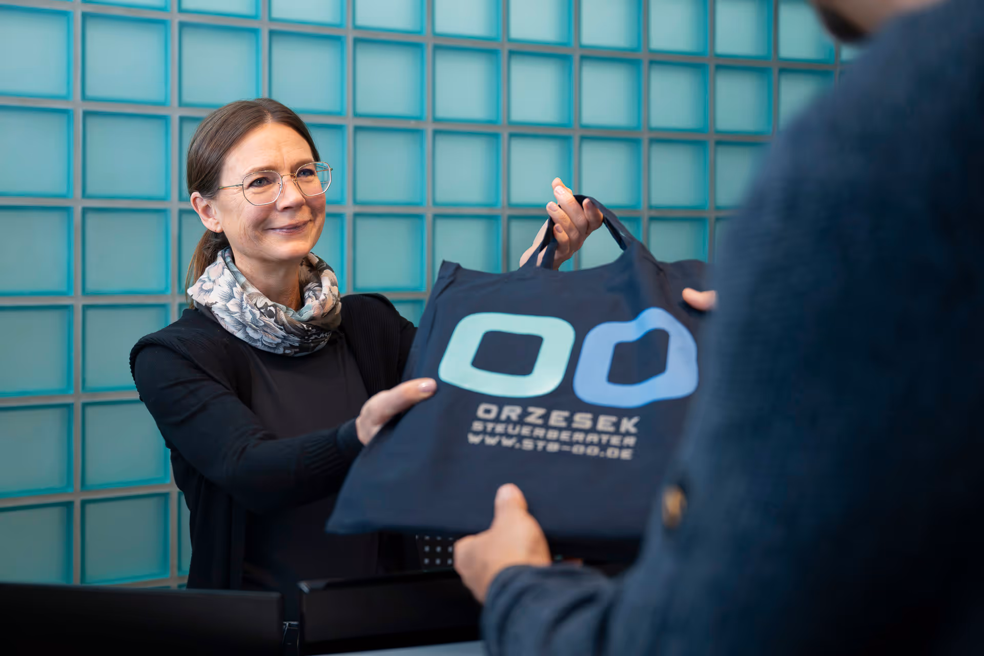 Eine Frau mit Brille und Schal übergibt eine Tasche mit der Aufschrift ORZESEK STEUERBERATER an eine andere Person.