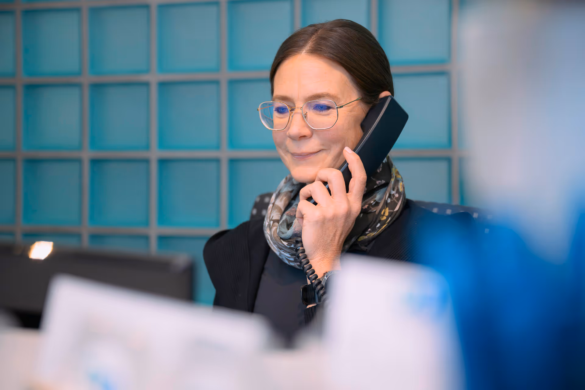 Frau mit Brille spricht am Telefon in einem Büro mit blauer Glaswand im Hintergrund.