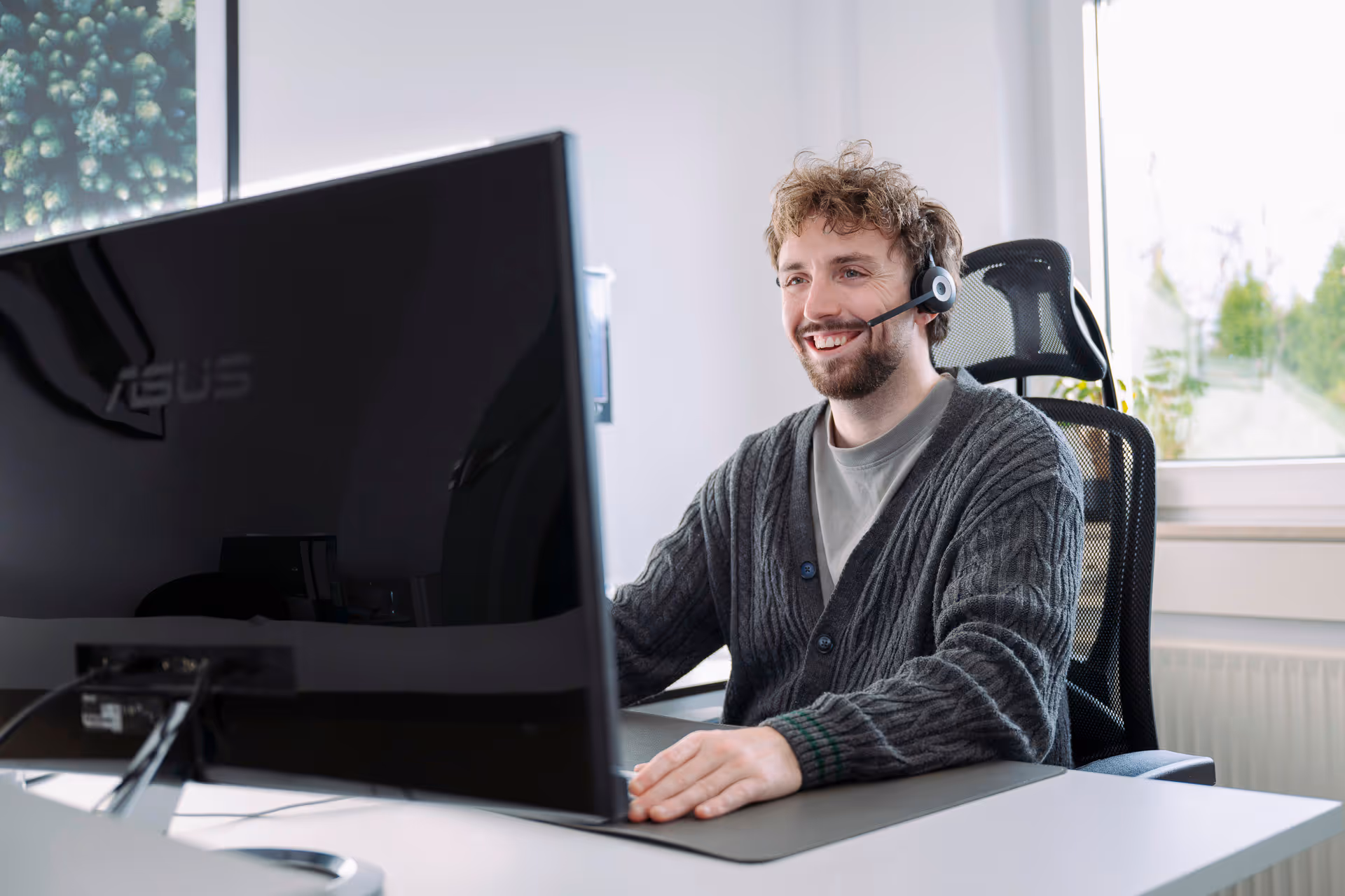 Junger Mann trägt Headset und lächelt, während er an einem Schreibtisch an einem Computer arbeitet.