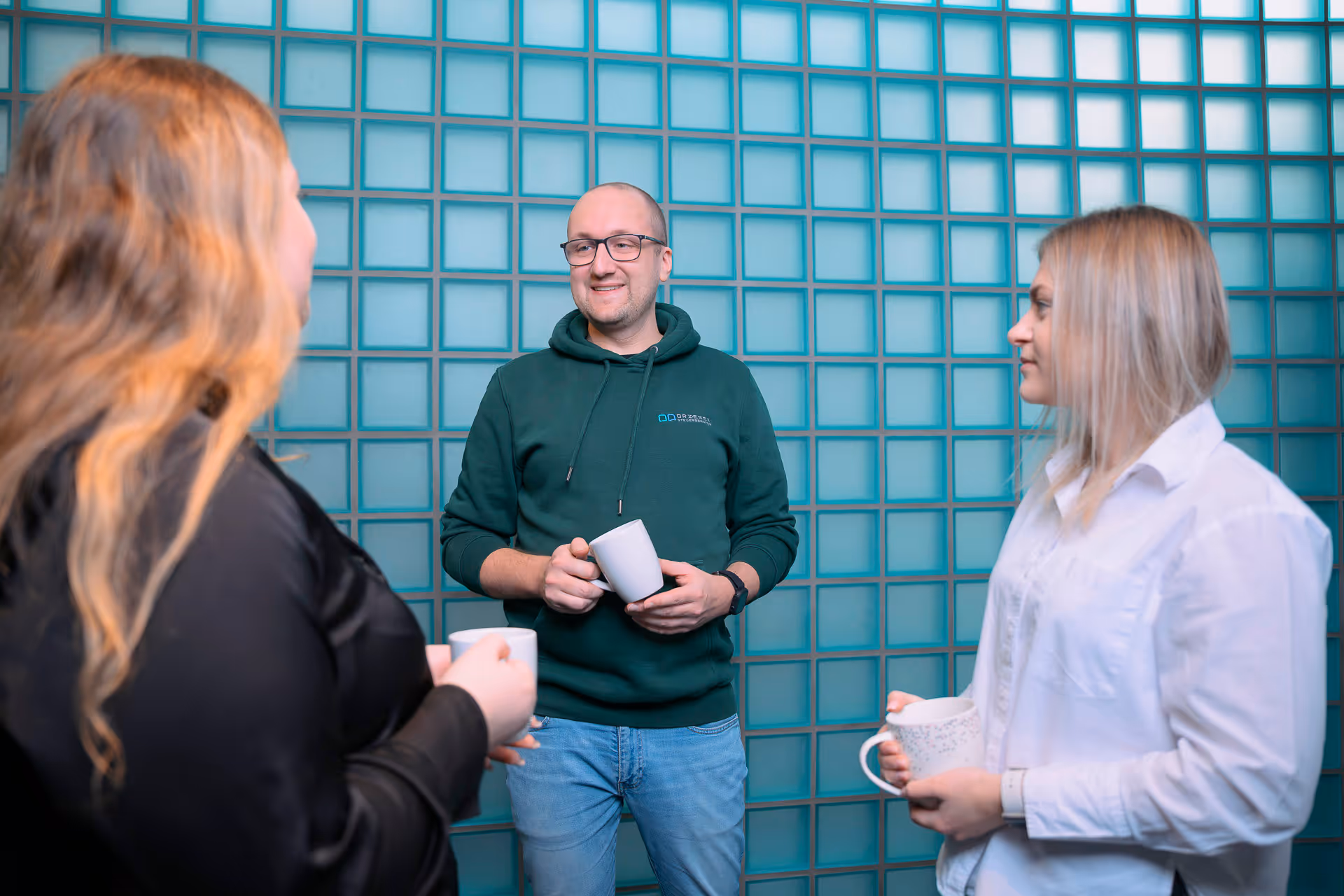 Drei Personen stehen vor einer Glasstein Wand und unterhalten sich, während sie Kaffeetassen halten.