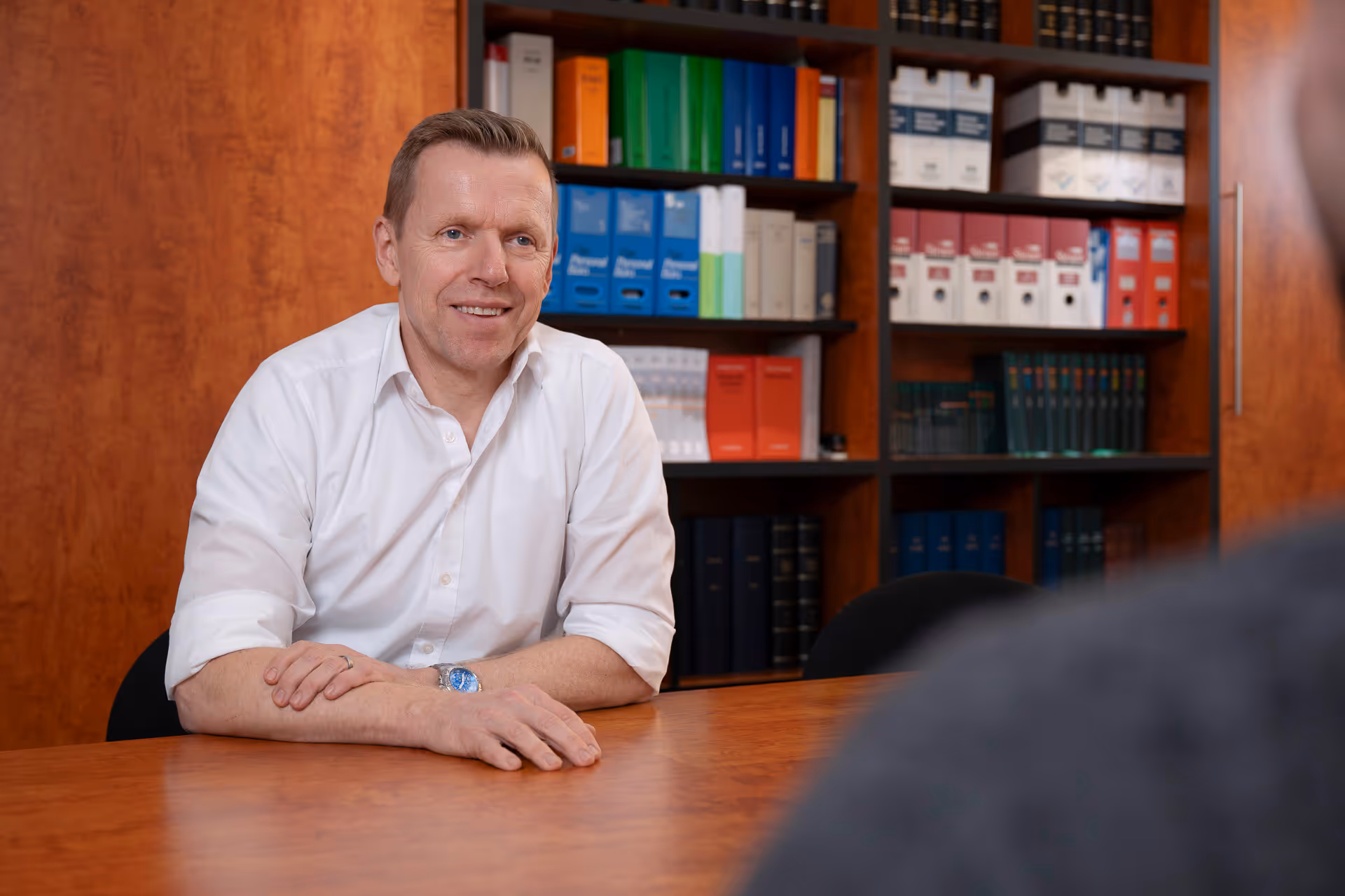 Michael Orzesek im weißen Hemd, der an einem Holztisch sitzt und lächelt, mit einem Bücherregal im Hintergrund.