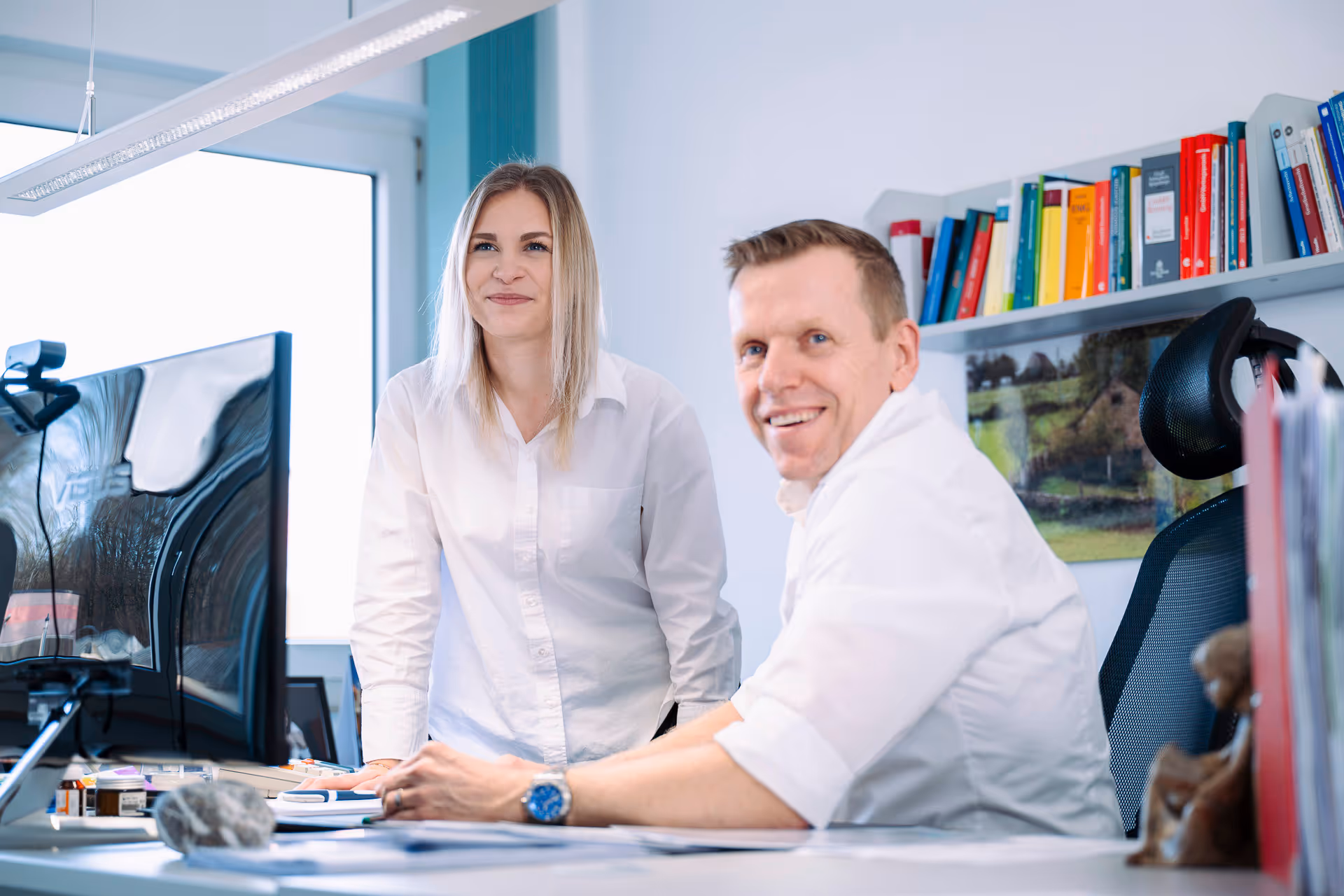 Zwei lächelnde Personen in einem Büro, eine Frau steht und ein Mann sitzt vor einem Computerbildschirm, umgeben von Büchern und Büroausstattung.