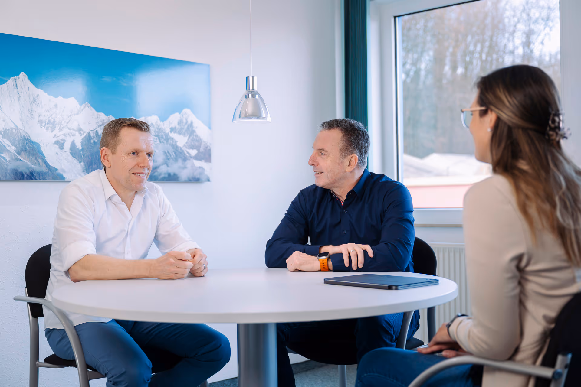 Drei Personen führen ein Gespräch an einem runden Tisch in einem hellen Raum mit einem Bild von verschneiten Bergen im Hintergrund.
