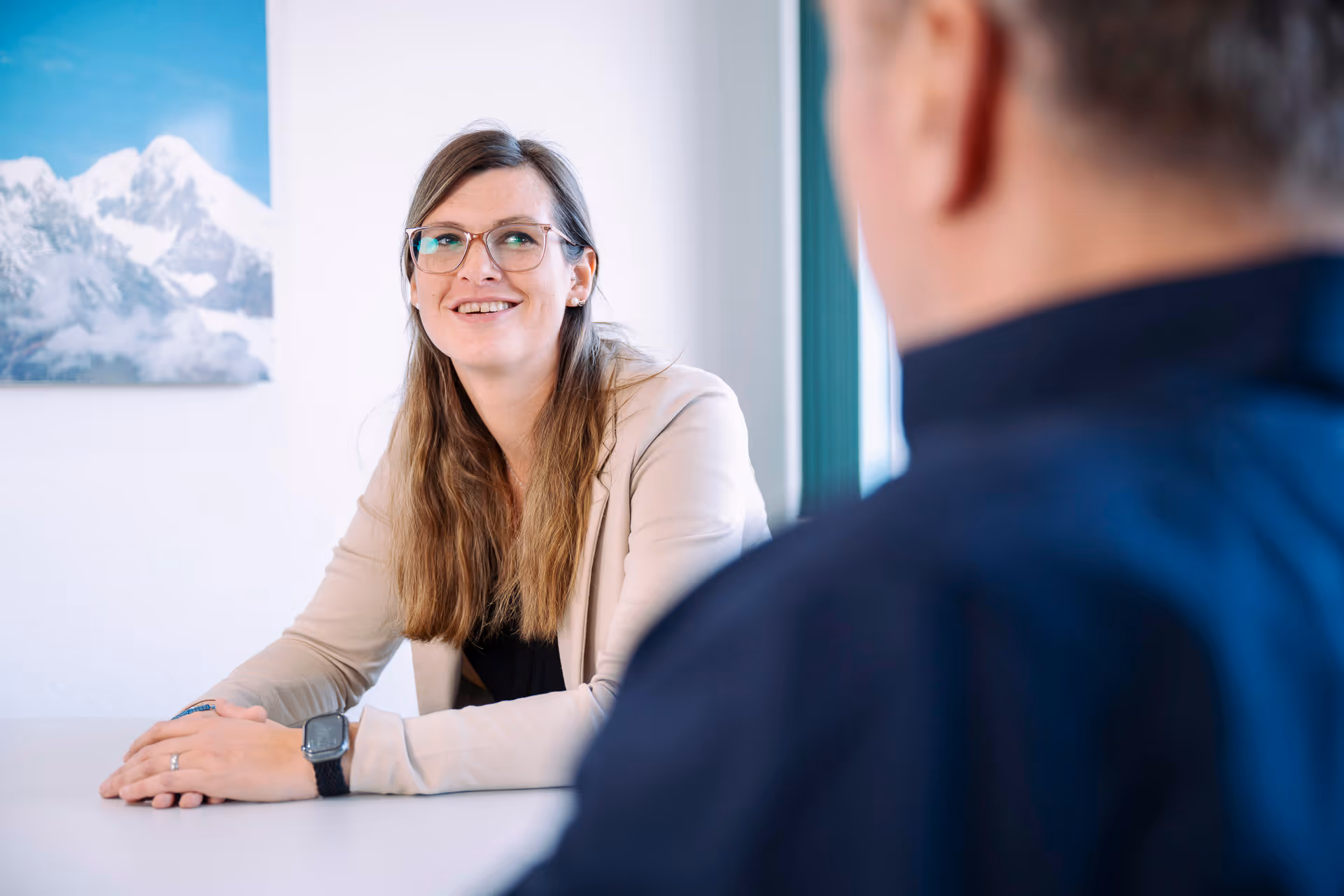 Frau mit Brille und beiger Jacke lächelt während eines Gesprächs mit einem Mann in einem Büro.