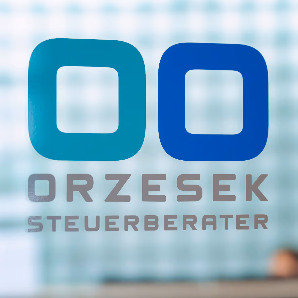 Logo von Orzesek Steuerberater mit zwei quadratischen, abgerundeten Symbolen in Türkis und Blau auf einer Glasscheibe.