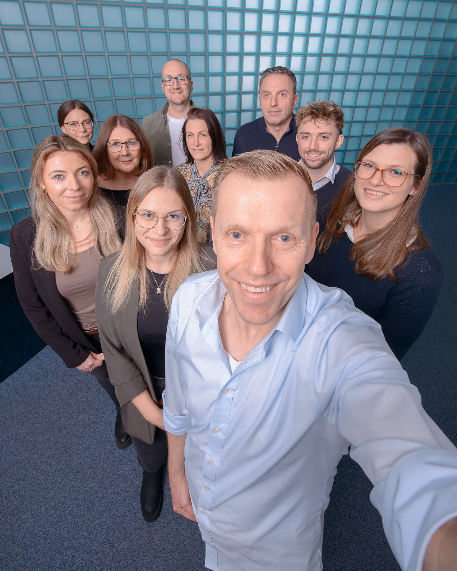 Orzesek Team von zehn Personen, die in einem Büro vor einer Glasblockwand in die Kamera lächeln.