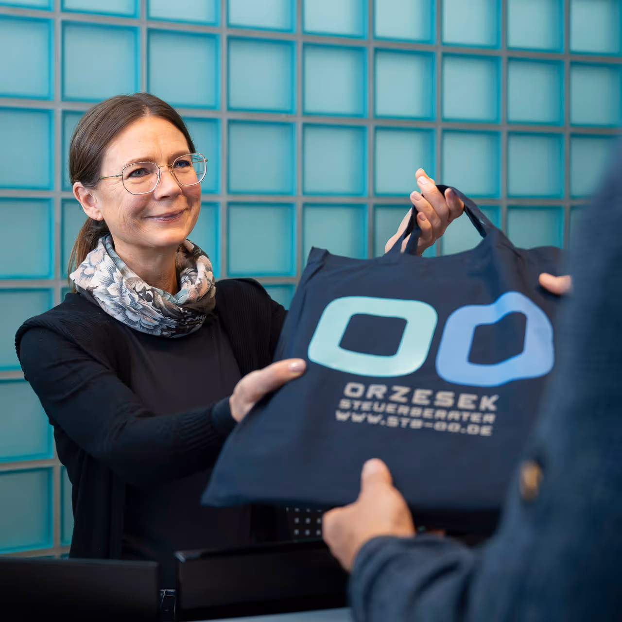Frau mit Brille und Schal übergibt eine Tasche mit dem Logo von Orzesek Steuerberater.