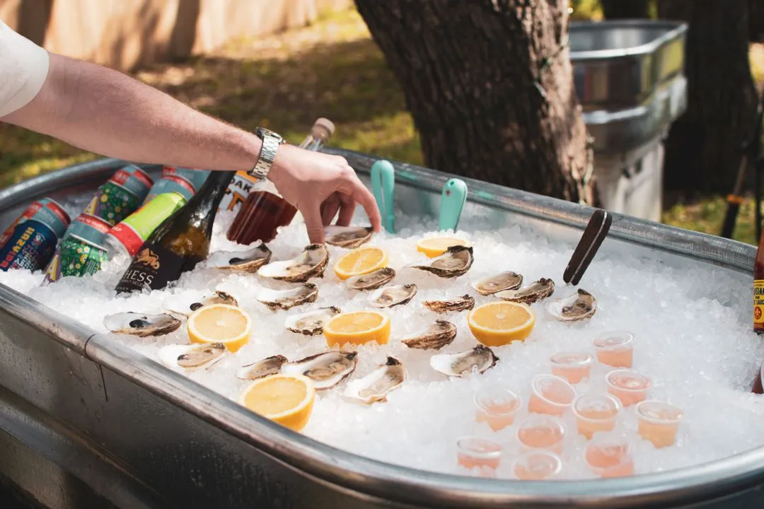 A close up of a person reaching into a trough with beverages and oysters on ice.