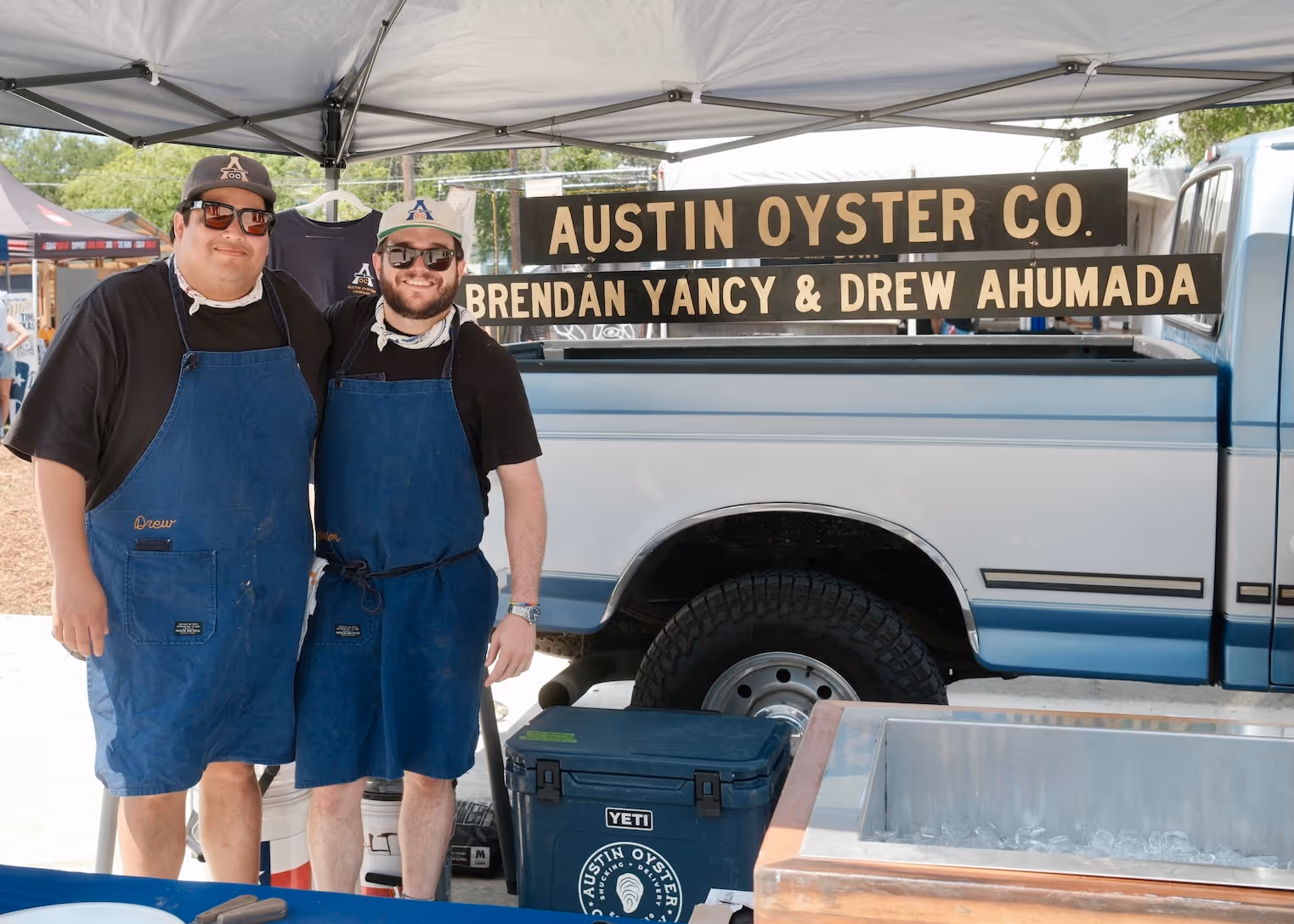 Two men wearing blue aprons and hats stand under a tent next to an Austin Oyster Co. sign and a pickup truck.