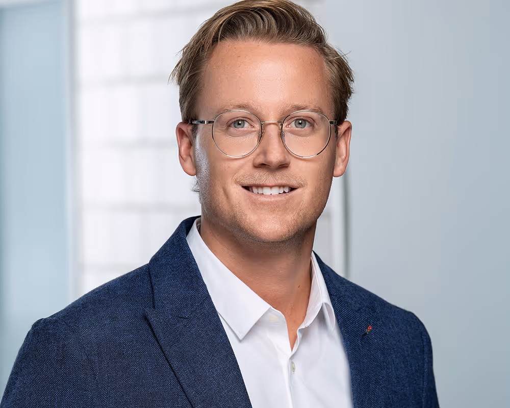Smiling young man with glasses wearing a white shirt and blue blazer against a light background.