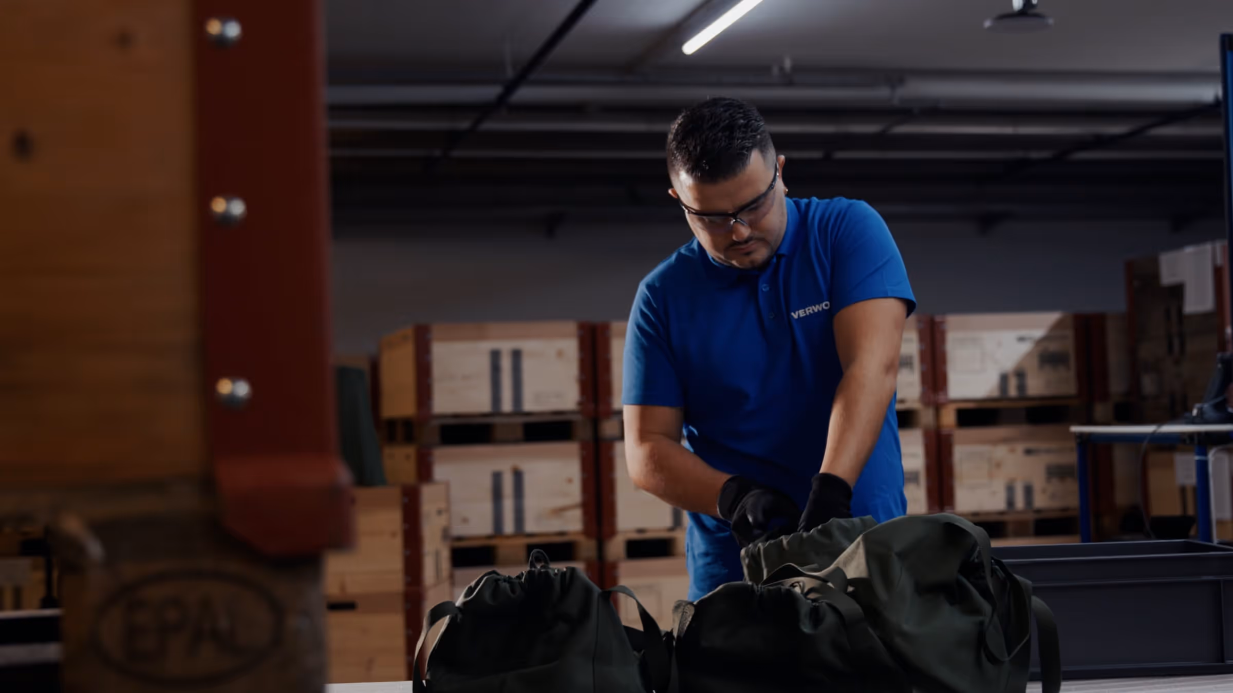 Man wearing safety glasses and gloves packing dark green bags in a warehouse with wooden crates stacked in the background.