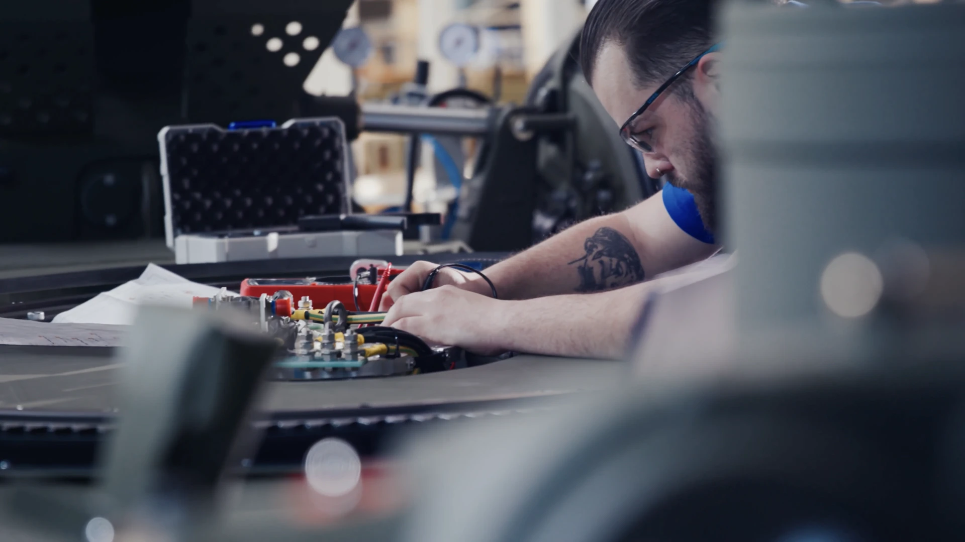 Technician with glasses working on electrical components with wires and tools in a workshop.