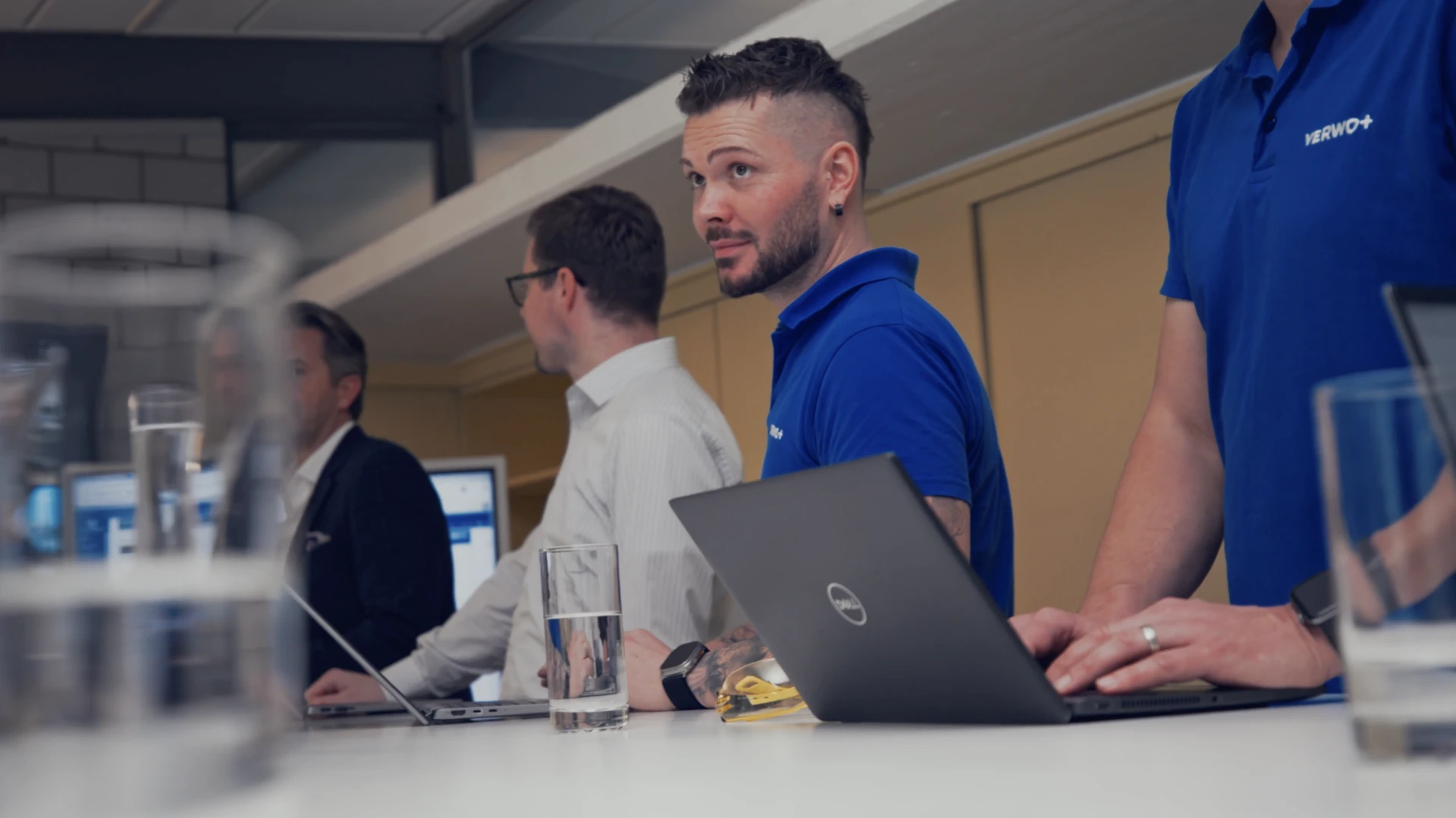 Three men working on laptops in an office, one wearing a blue polo shirt with Verwo+ logo.