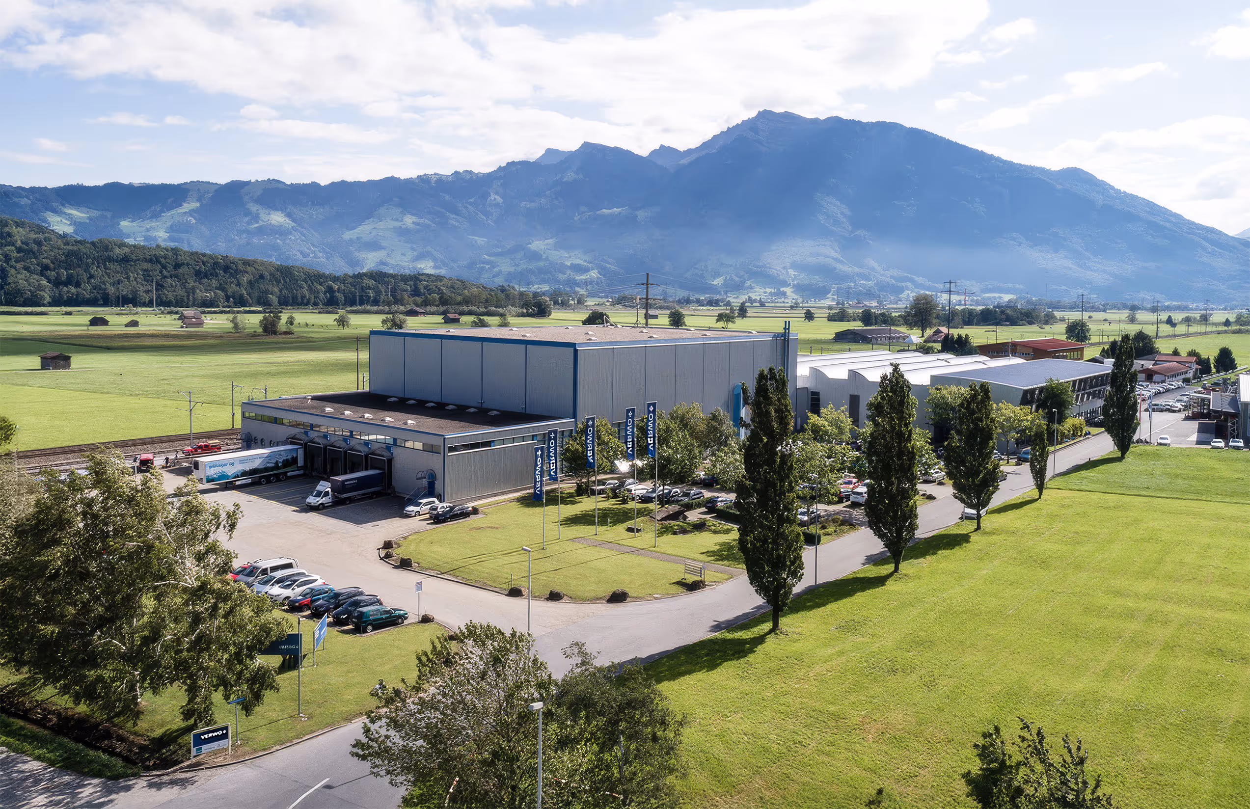 Aerial view of an industrial facility with parked cars, surrounded by green fields and mountains in the background under a partly cloudy sky.