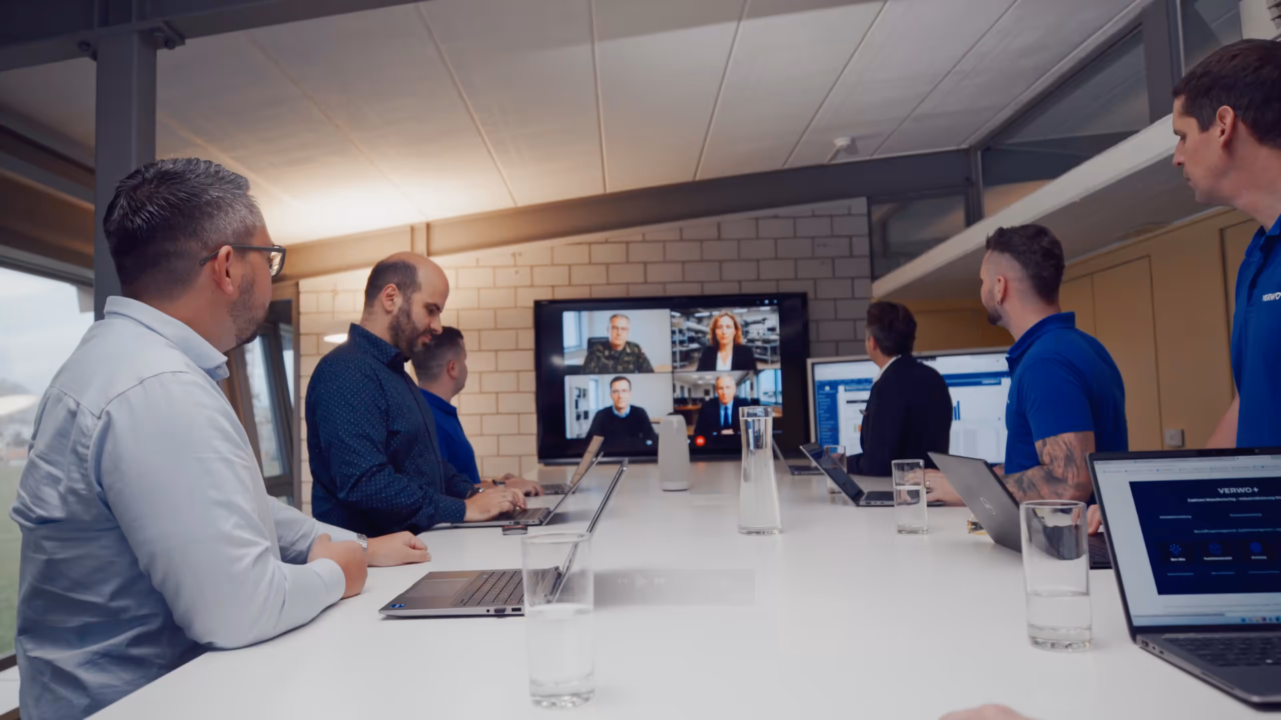 A group of men sitting around a conference table engaged in a video call with four people displayed on a large screen.