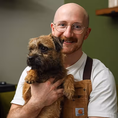 Smiling bald man, Craig, with glasses wearing an apron holding his small brown and black border terrier bog.