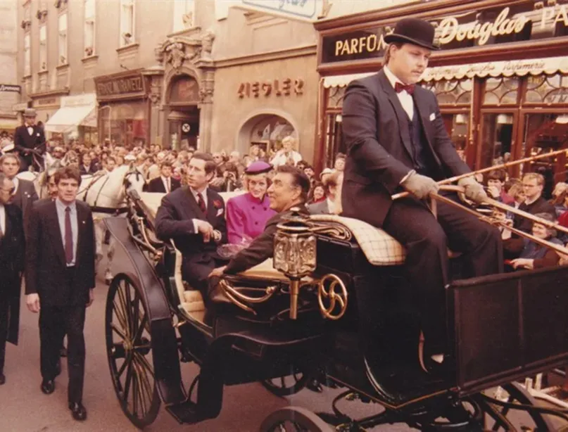Eine historische Pferdekutsche, gezogen von weißen Pferden, fährt durch eine belebte Stadtstraße mit vielen Zuschauern. Eine Frau in einem auffälligen pinken Mantel und Hut sitzt in der Kutsche.