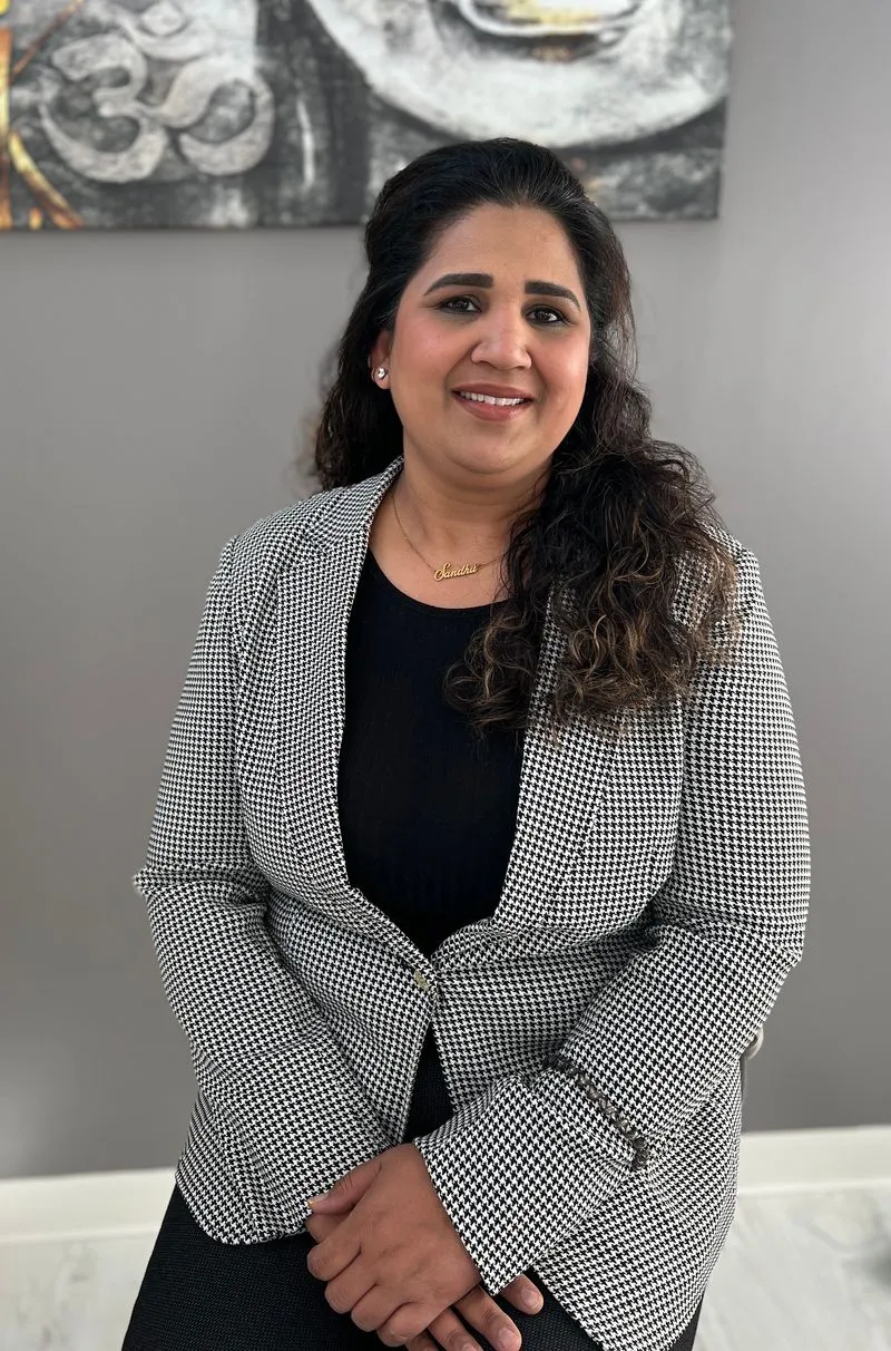Smiling woman with long dark hair wearing a black top and a black and white houndstooth blazer sitting with hands folded.